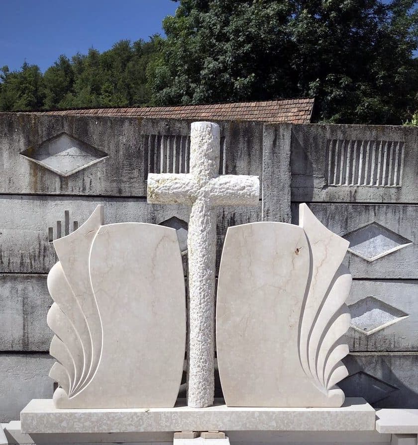 Monument funerar dublu din travertin crem cu cruce texturată și aripi sculptate, în atelierul Mogut.