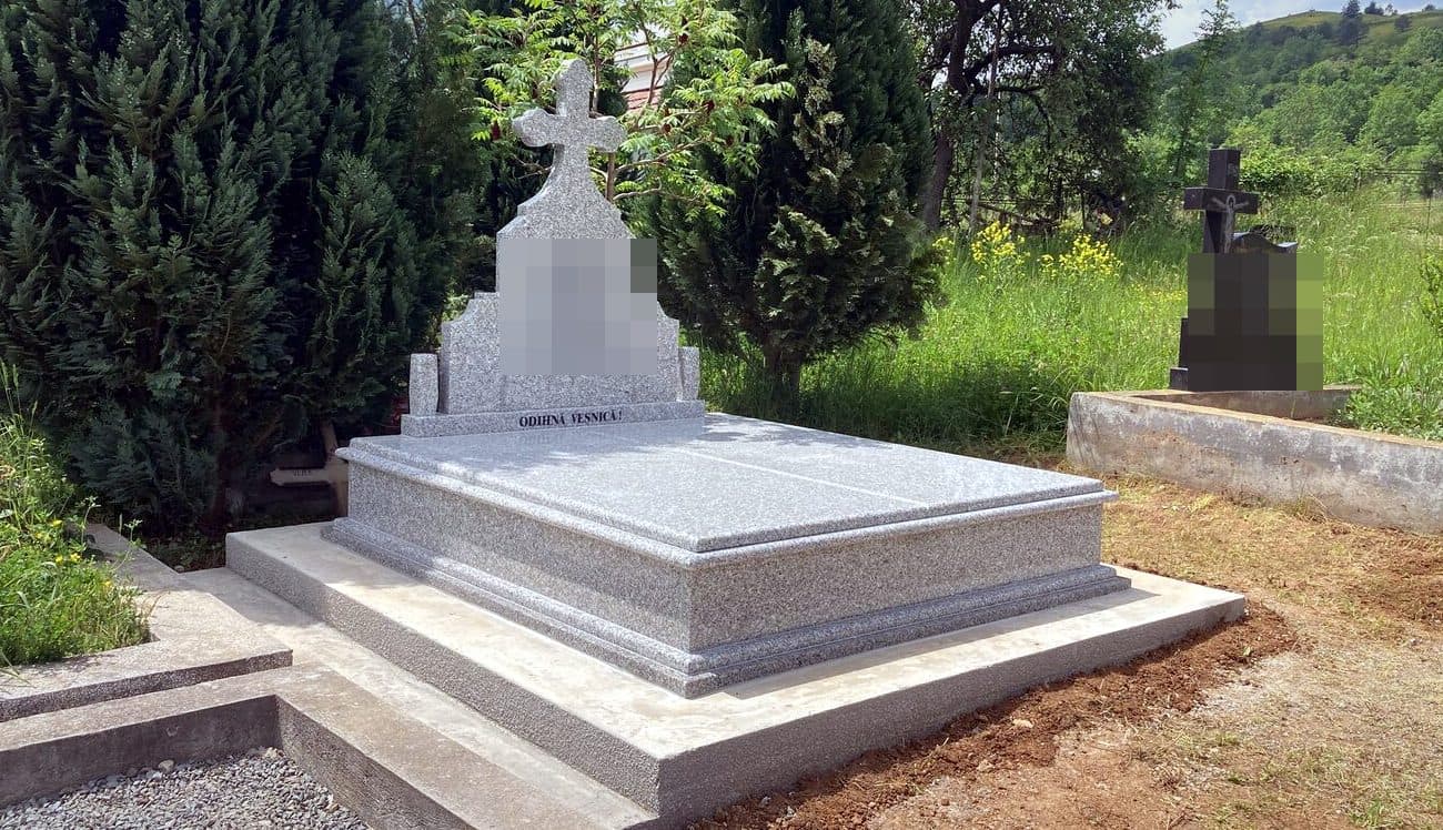 Mormânt clasic din granit gri cu cruce, montat în cimitir într-un cadru verde.