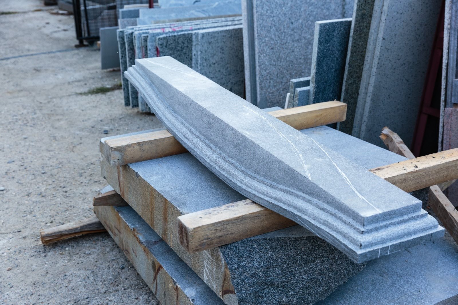 Piesă de granit gri profilată în trepte și curbată, așezată pe șipci de lemn în curtea atelierului, cu plăci de granit în fundal